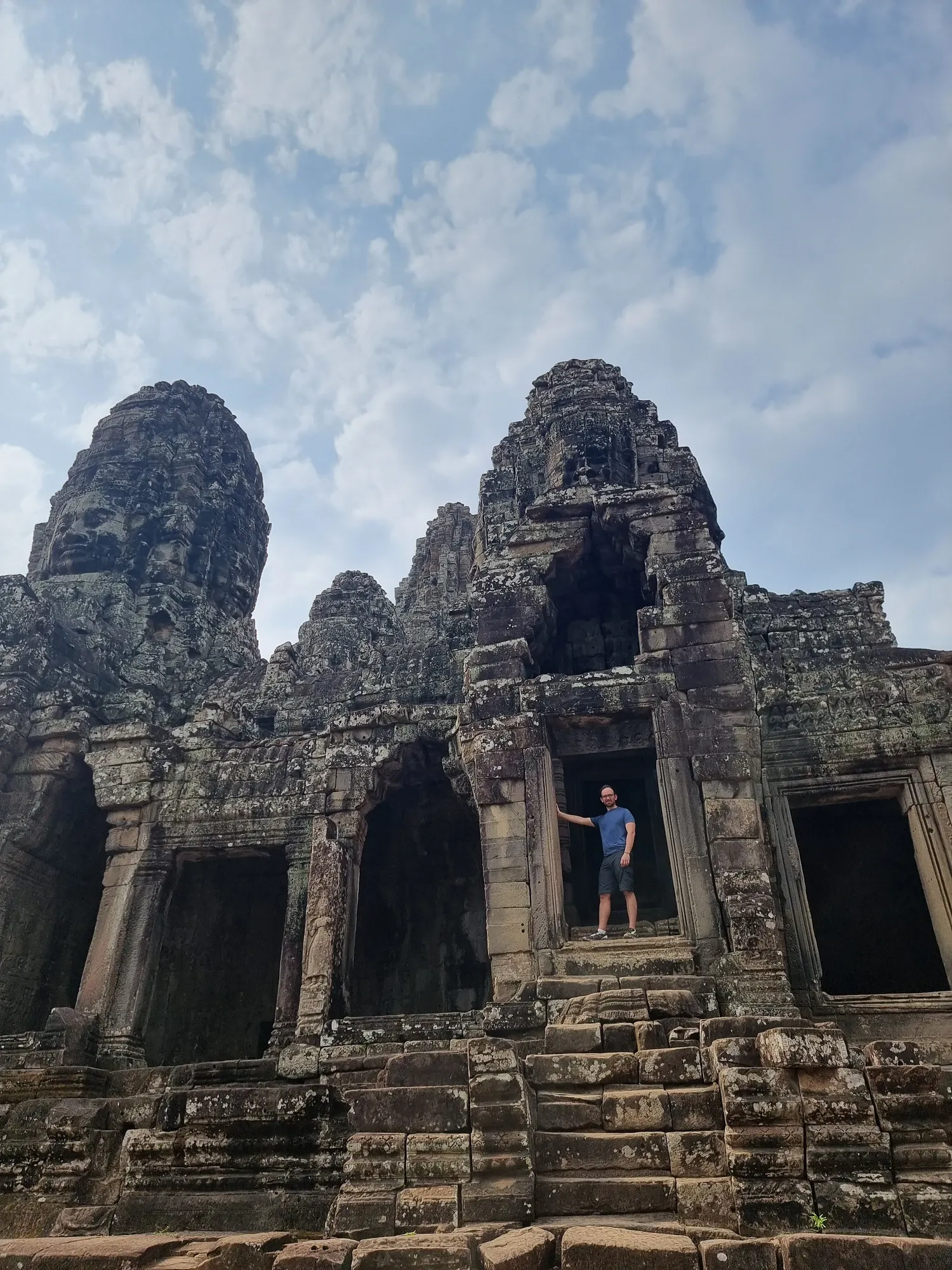 Antoine, en t-shirt bleu, debout dans une porte du Bayon, sous deux tours sculptées de visages