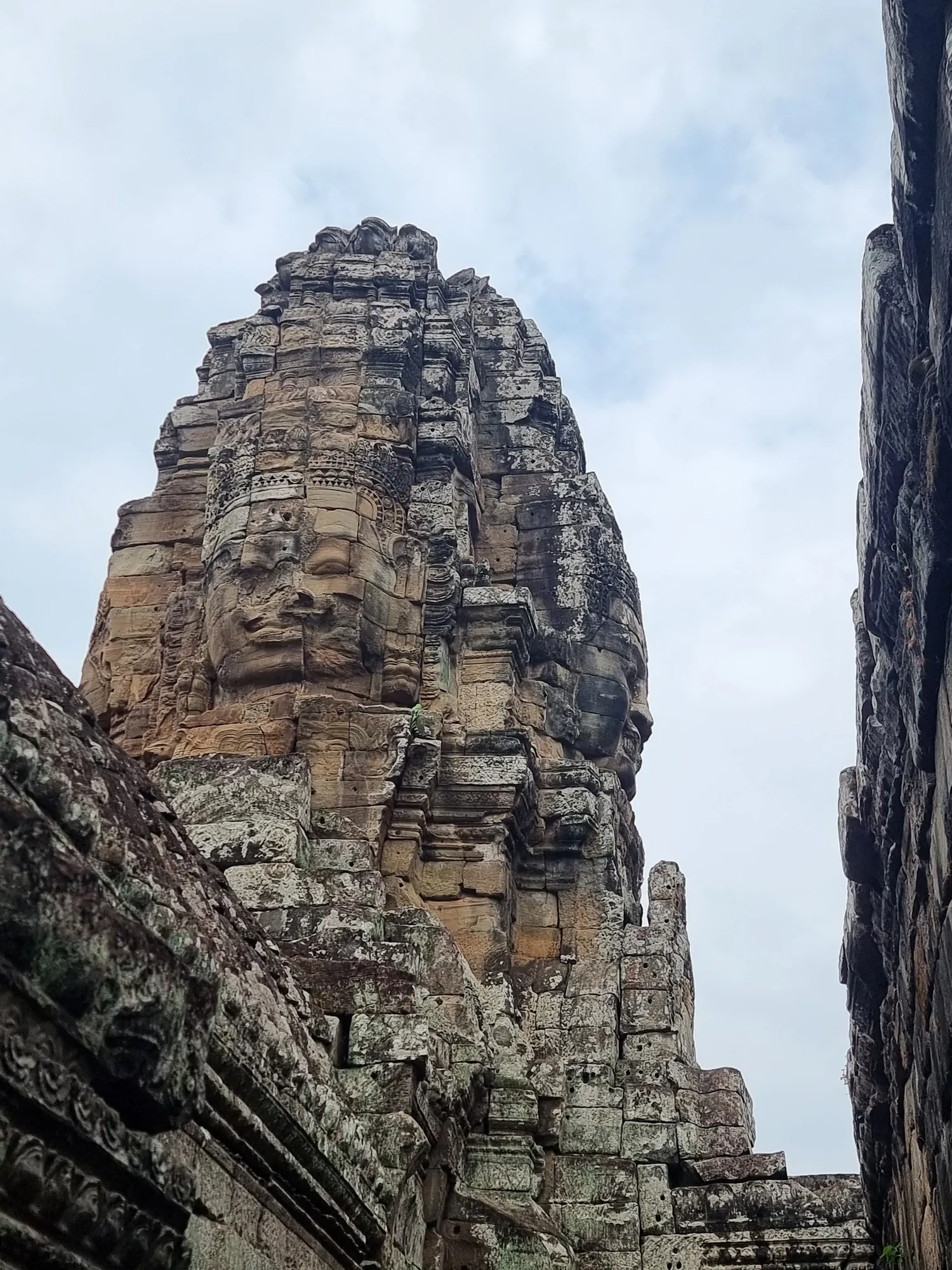 Un visage sculpté géant sur une tour du Bayon, vu en contre-plongée, sous un ciel nuageux