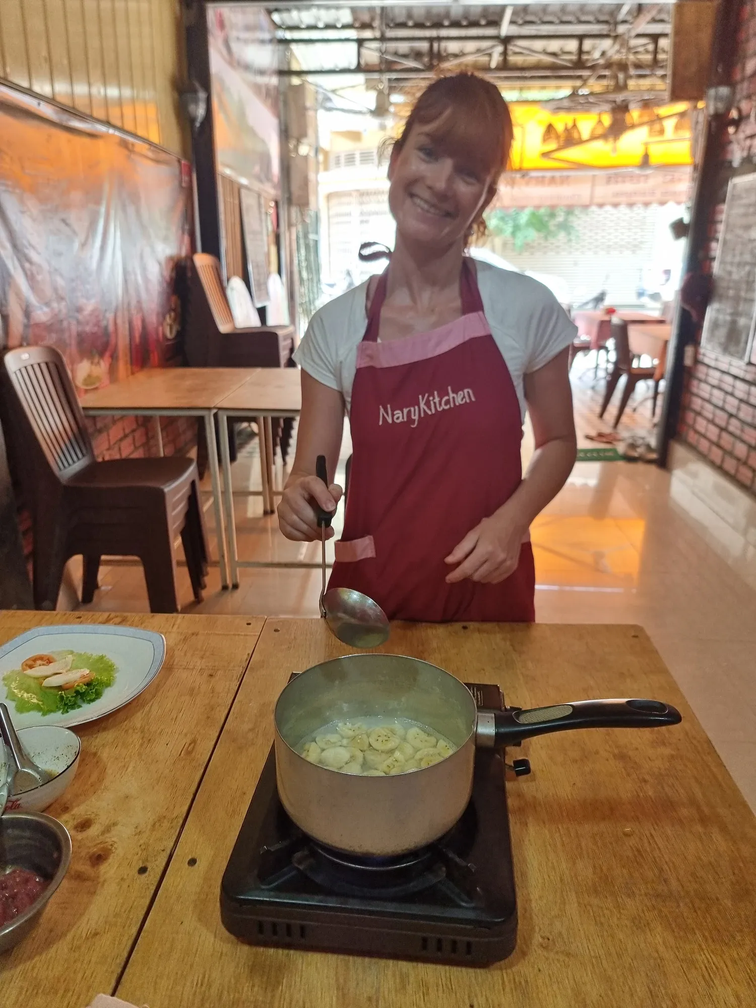 Ségolène, souriante, en tablier Nary Kitchen, une louche à la main devant une casserole posée sur un réchaud