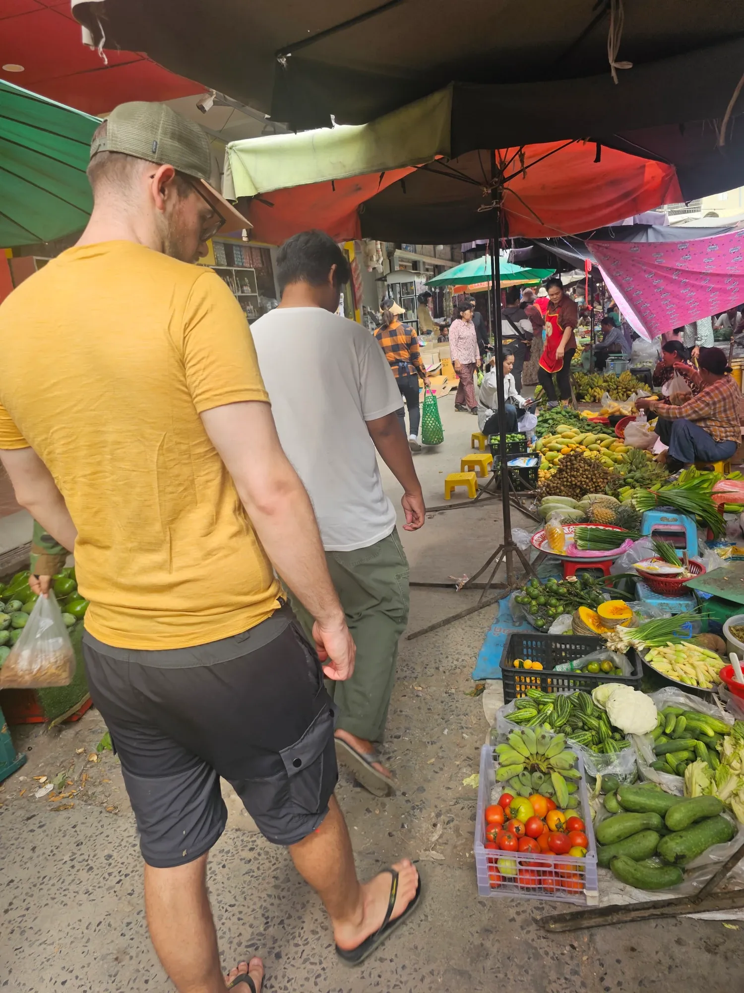 Antoine, de dos, en t-shirt jaune et casquette, dans une allée de marché bordée d'étals de légumes, de piments et de concombres