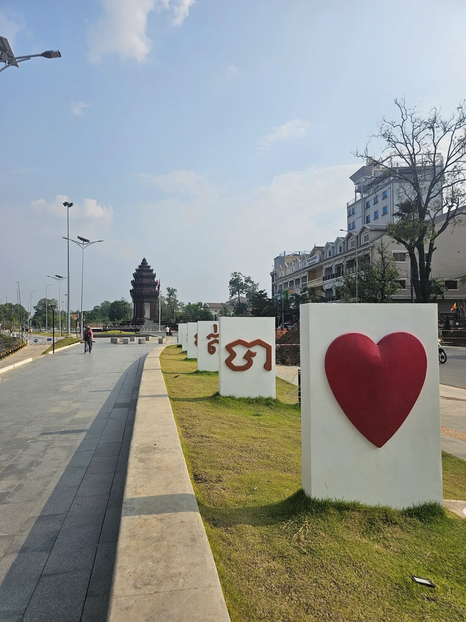 Promenade le long des quais de Battambang, bordée de blocs blancs ornés d'un cœur rouge et de lettres khmères, avec une tour de monument au loin sous un ciel bleu