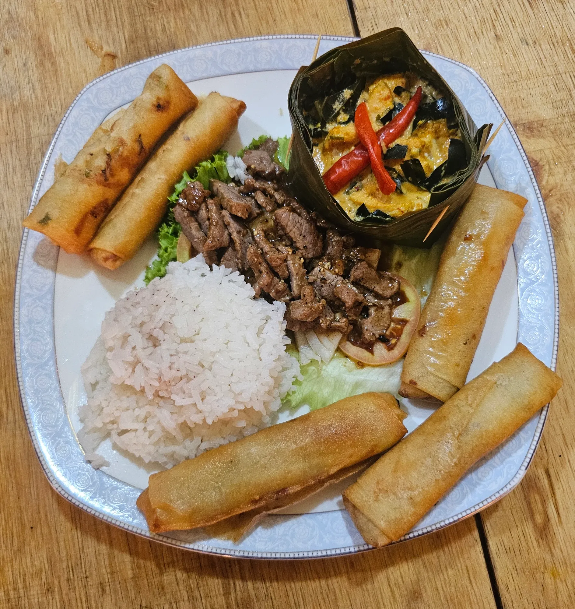 Assiette avec quatre nems dorés, un amok dans sa feuille de bananier, du bœuf lok lak sur de la salade et du riz blanc, sur une table en bois
