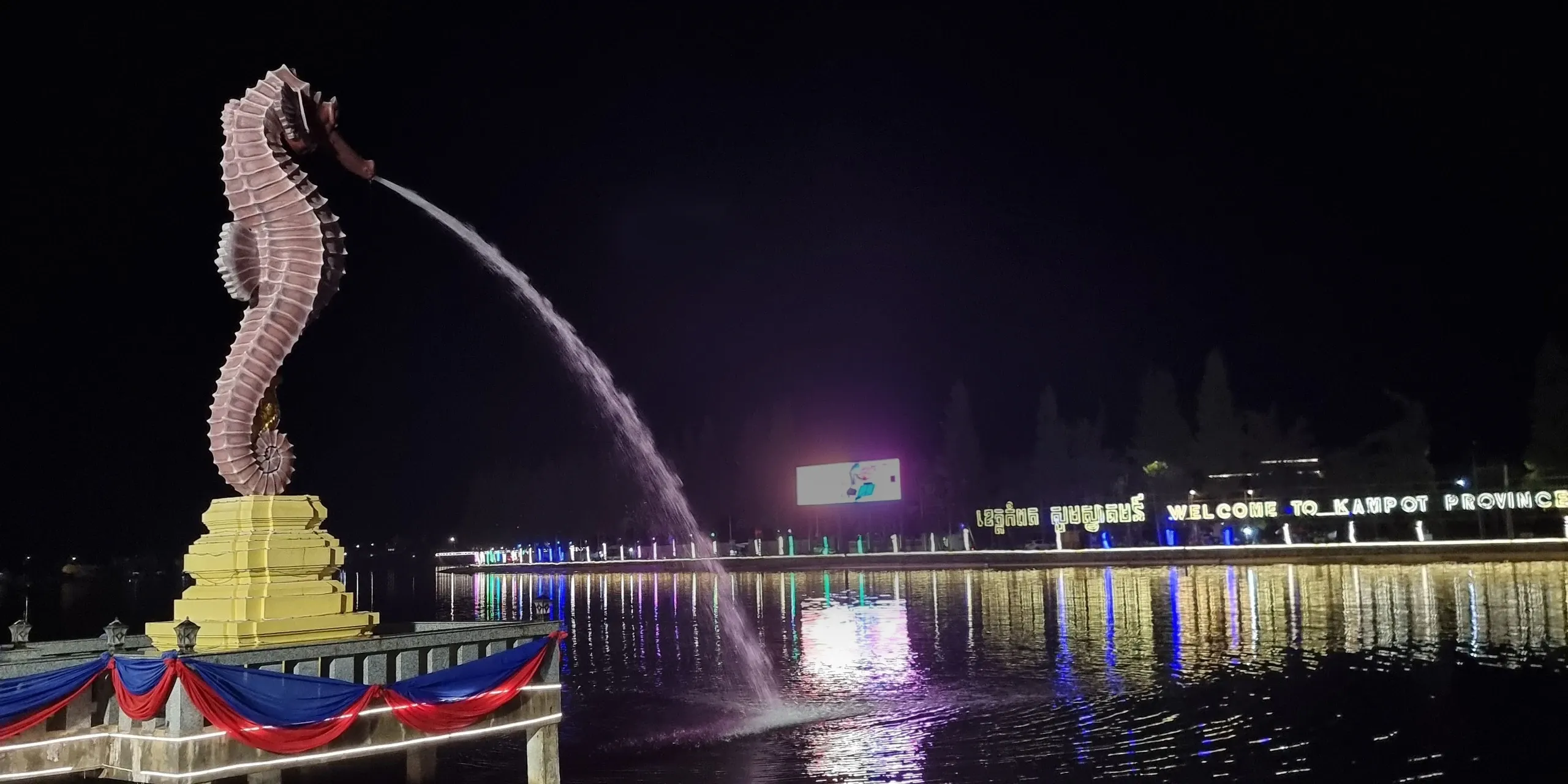 Statue géante d'hippocampe illuminée de nuit à Kampot, qui crache un jet d'eau, face à l'enseigne lumineuse « Welcome to Kampot Province », au-dessus des reflets des lumières sur l'eau