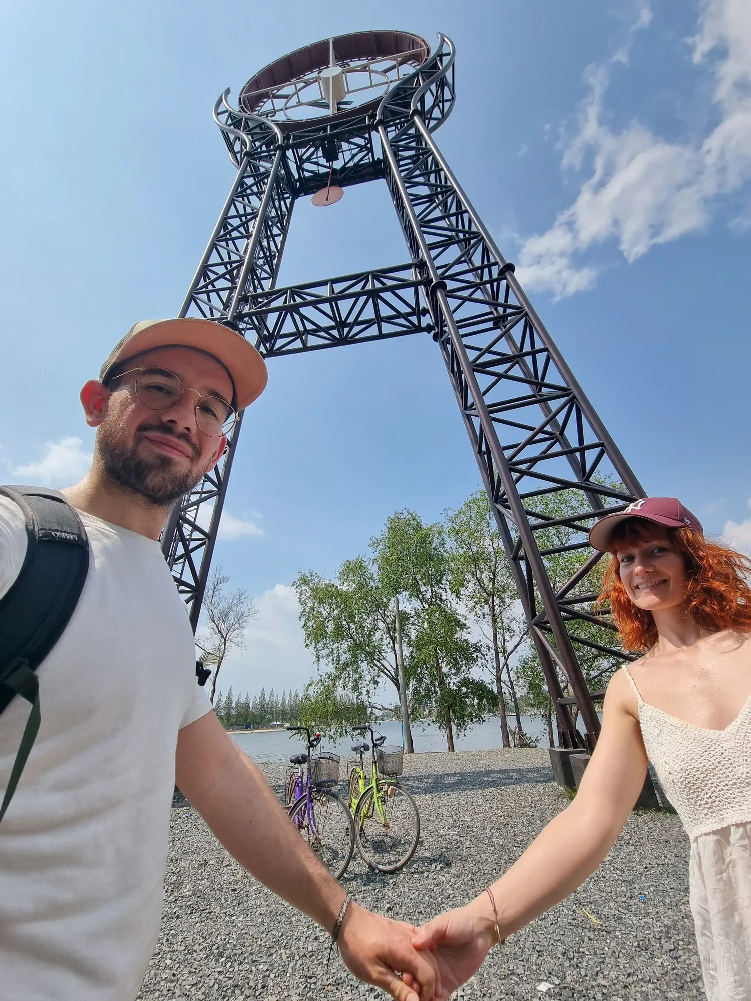 Antoine et Ségolène main dans la main, en selfie au pied de la grande horloge métallique de Kampot, leurs deux vélos garés derrière eux au bord de l'eau