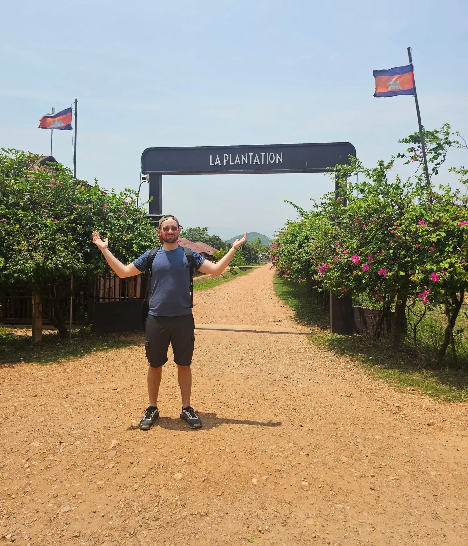 Antoine, les bras ouverts, devant le portique d'entrée de La Plantation, entre deux drapeaux cambodgiens et des massifs de fleurs roses, au début d'un chemin de terre