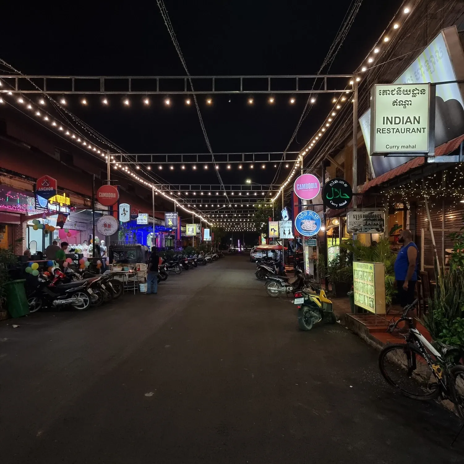 Une rue de Kampot le soir sous des guirlandes lumineuses, bordée de scooters garés et d'enseignes, dont celle du restaurant indien Curry Mahal à droite