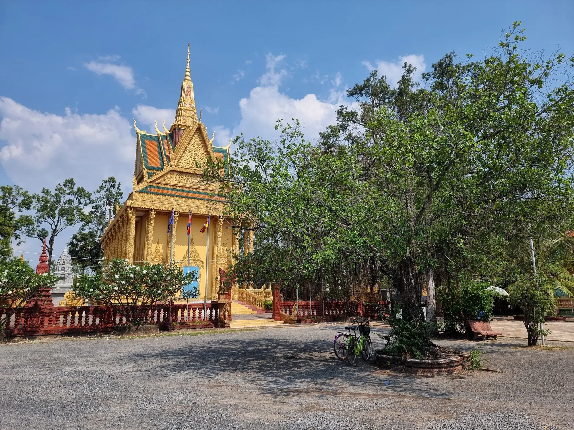 Deux vélos garés à l'ombre d'un arbre devant le bâtiment doré du temple Traeuy Kaoh, sous un ciel bleu
