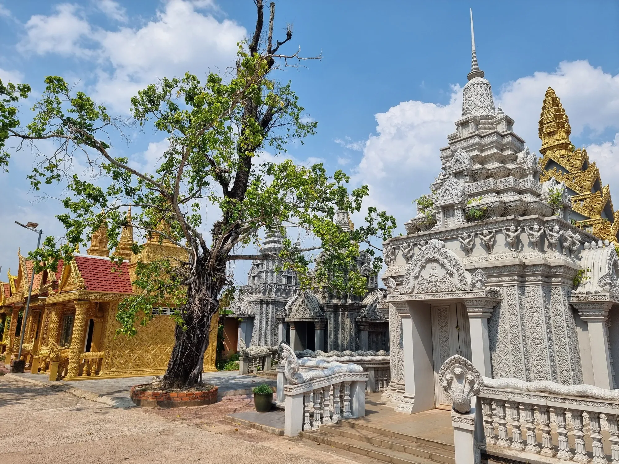 Le temple Traeuy Kaoh, avec ses édifices dorés et ses tours blanches sculptées autour d'un grand arbre, sous un ciel bleu