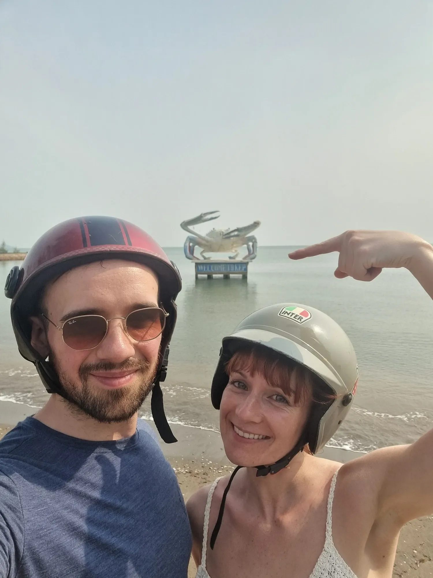 Antoine et Ségolène en selfie sur la plage de Kep, casques de scooter sur la tête, Ségolène pointant du doigt la statue du crabe dans la mer derrière eux