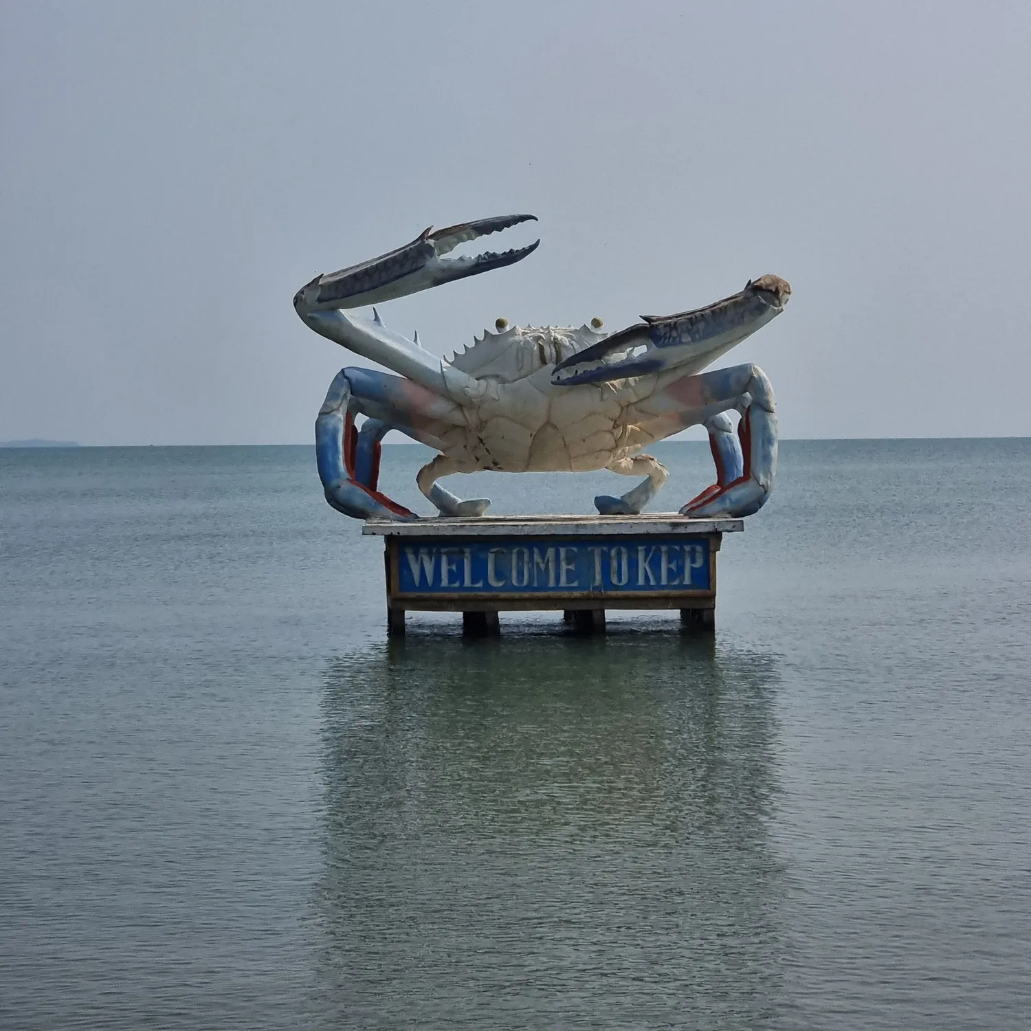 La statue géante d'un crabe bleu et blanc dans la mer, sur un socle « Welcome to Kep »
