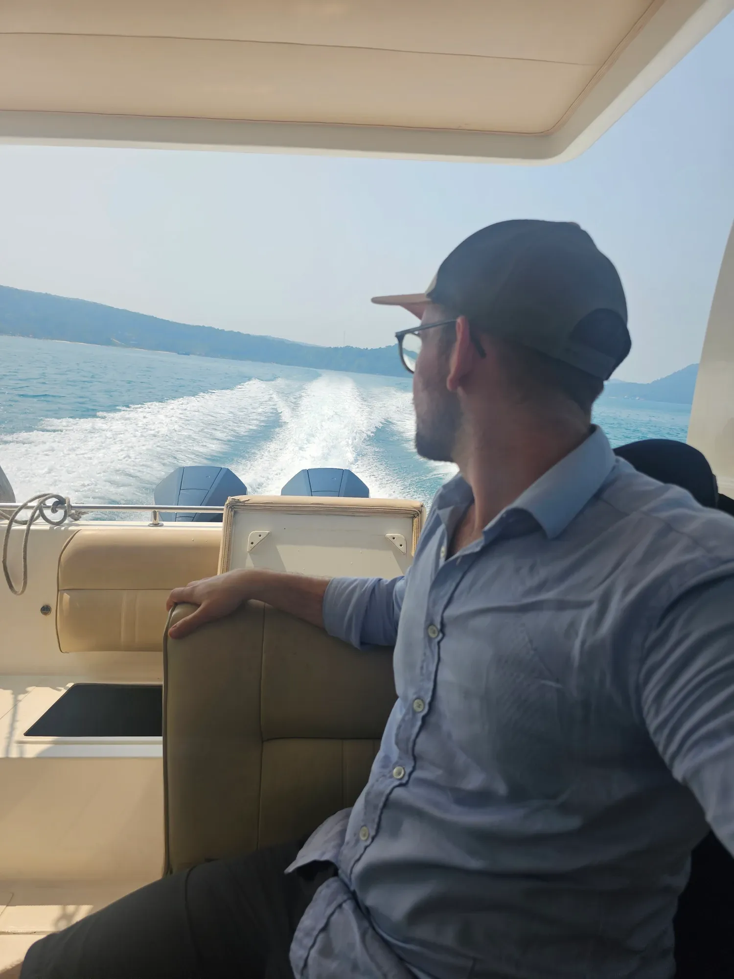 Antoine, de profil, casquette et chemise bleue, assis à l'arrière d'un bateau à moteur, avec derrière lui le sillage blanc sur la mer et une côte boisée