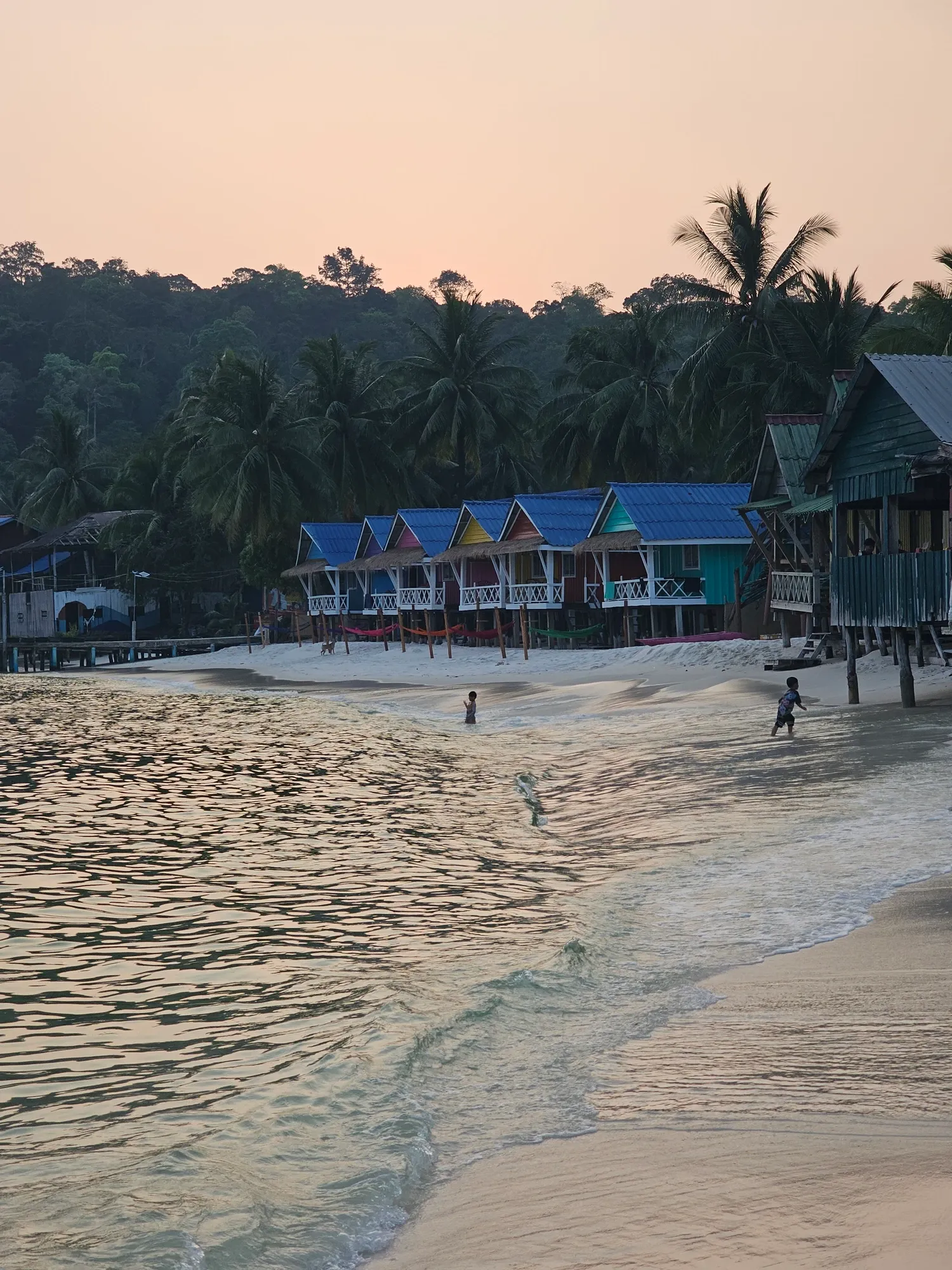 Rangée de bungalows colorés sur pilotis au bord d'une plage, sous un ciel rose de fin de journée, avec des palmiers et la forêt derrière, et deux enfants qui jouent dans les vagues