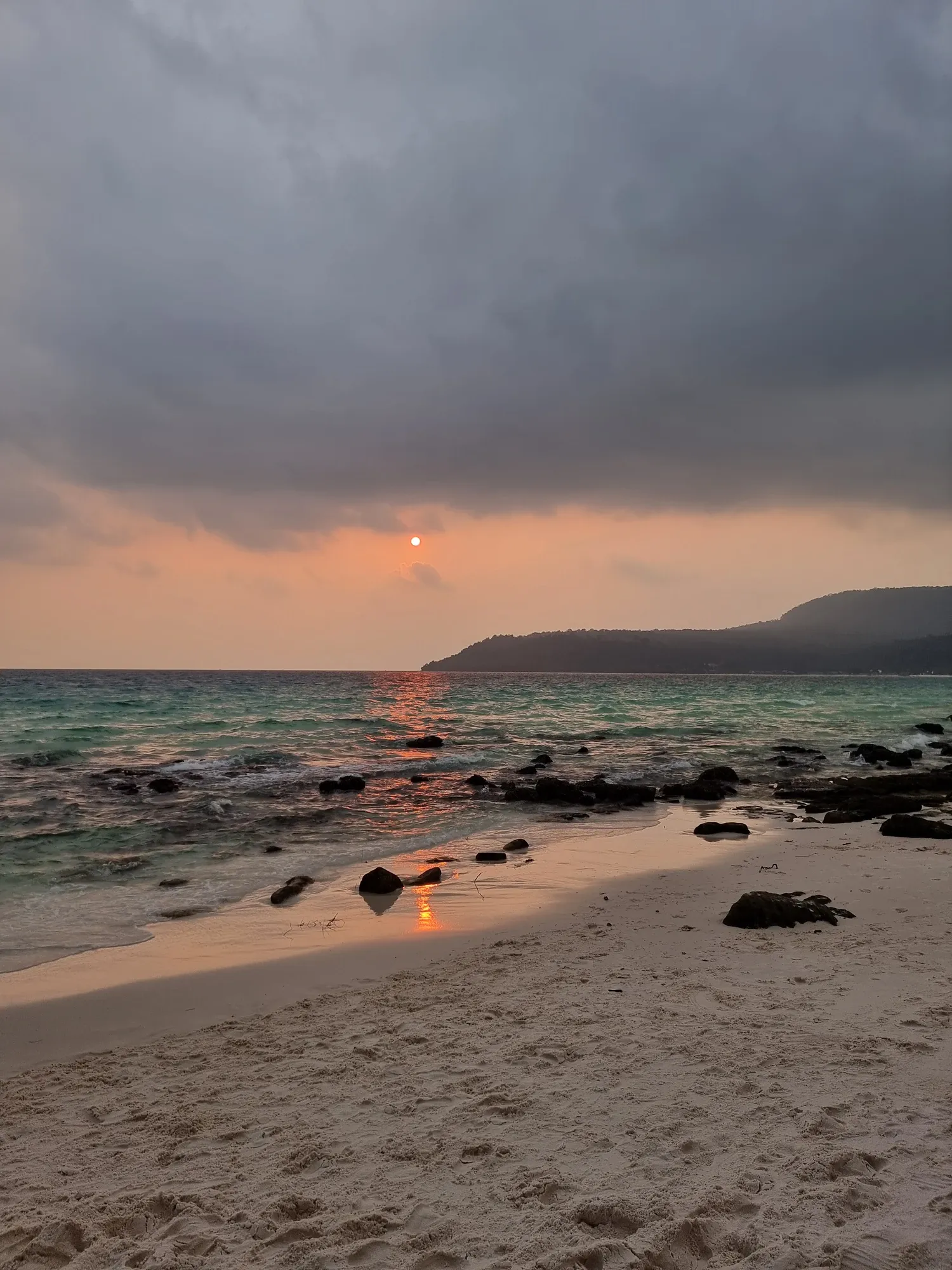 Coucher de soleil sous un ciel nuageux, le soleil se reflète sur la mer et sur le sable mouillé entre des rochers sombres, avec une colline boisée à droite