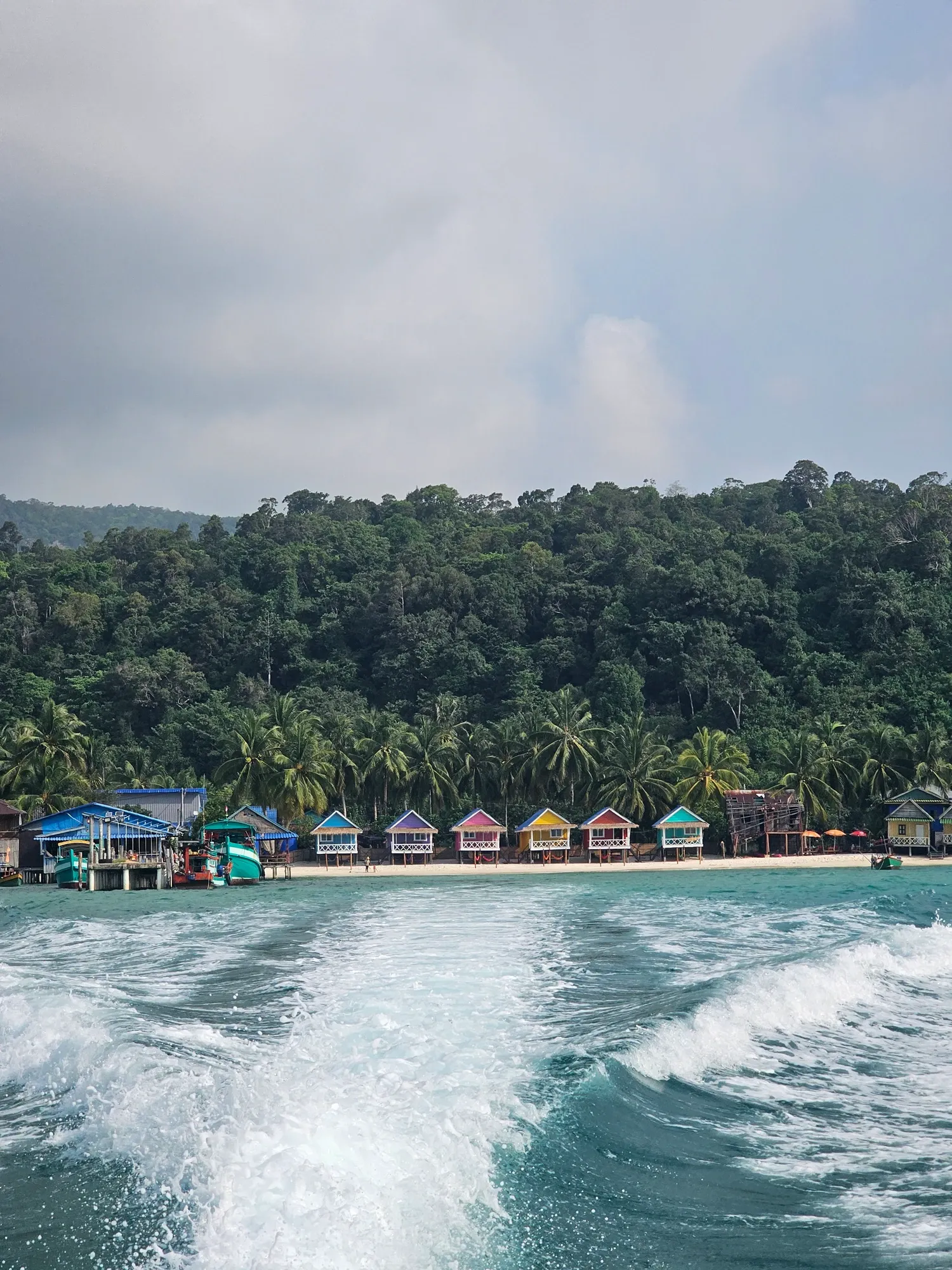 Le sillage blanc d'un bateau sur la mer, et au loin la rangée de bungalows colorés au pied d'une colline couverte de forêt, sous un ciel gris
