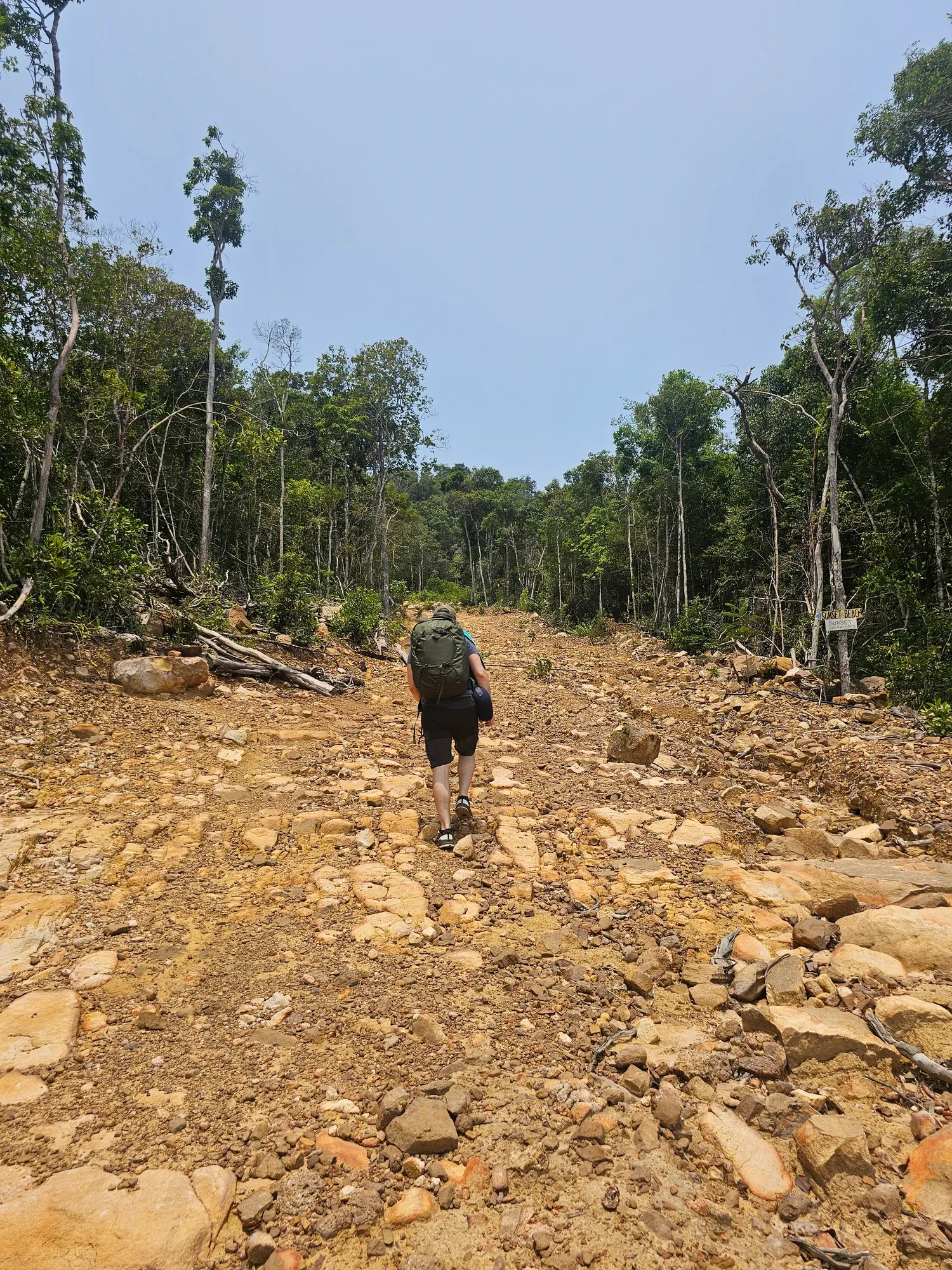 Antoine, de dos avec son sac à dos, gravit une large piste de terre et de cailloux bordée de jungle, à côté d'un panneau indiquant Sunset Beach