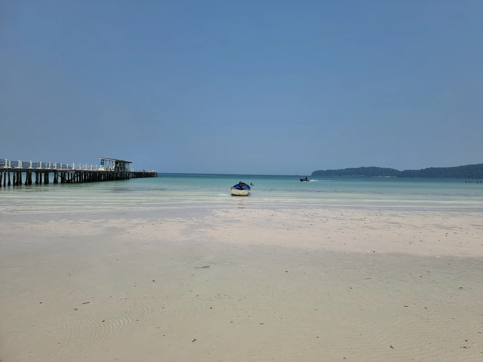 Plage de sable blanc de Saracen Bay, avec un long ponton en bois à gauche, une petite barque dans l'eau turquoise et une côte boisée à l'horizon
