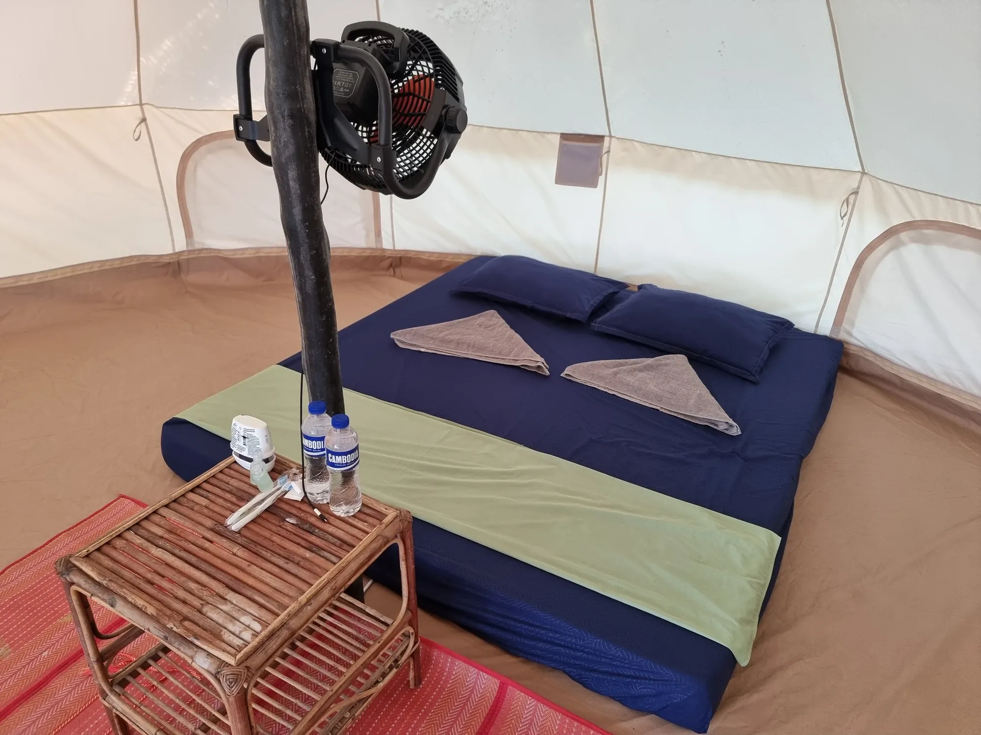 Intérieur de notre tipi au Sleeping Trees : un matelas aux draps bleus avec deux serviettes pliées, un ventilateur fixé au mât central et une petite table en bambou avec deux bouteilles d'eau