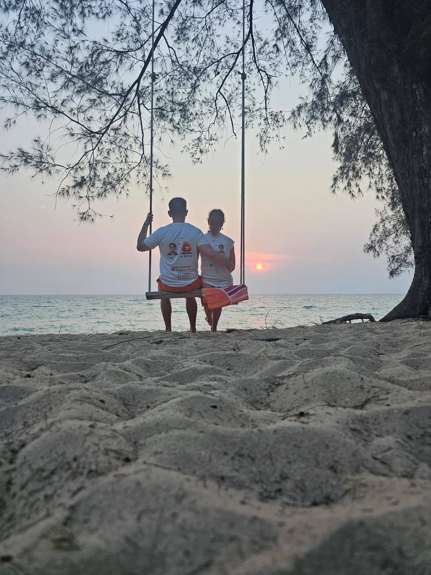 Antoine et Ségolène, de dos, assis côte à côte sur une balançoire suspendue à un arbre, face au soleil couchant sur la mer