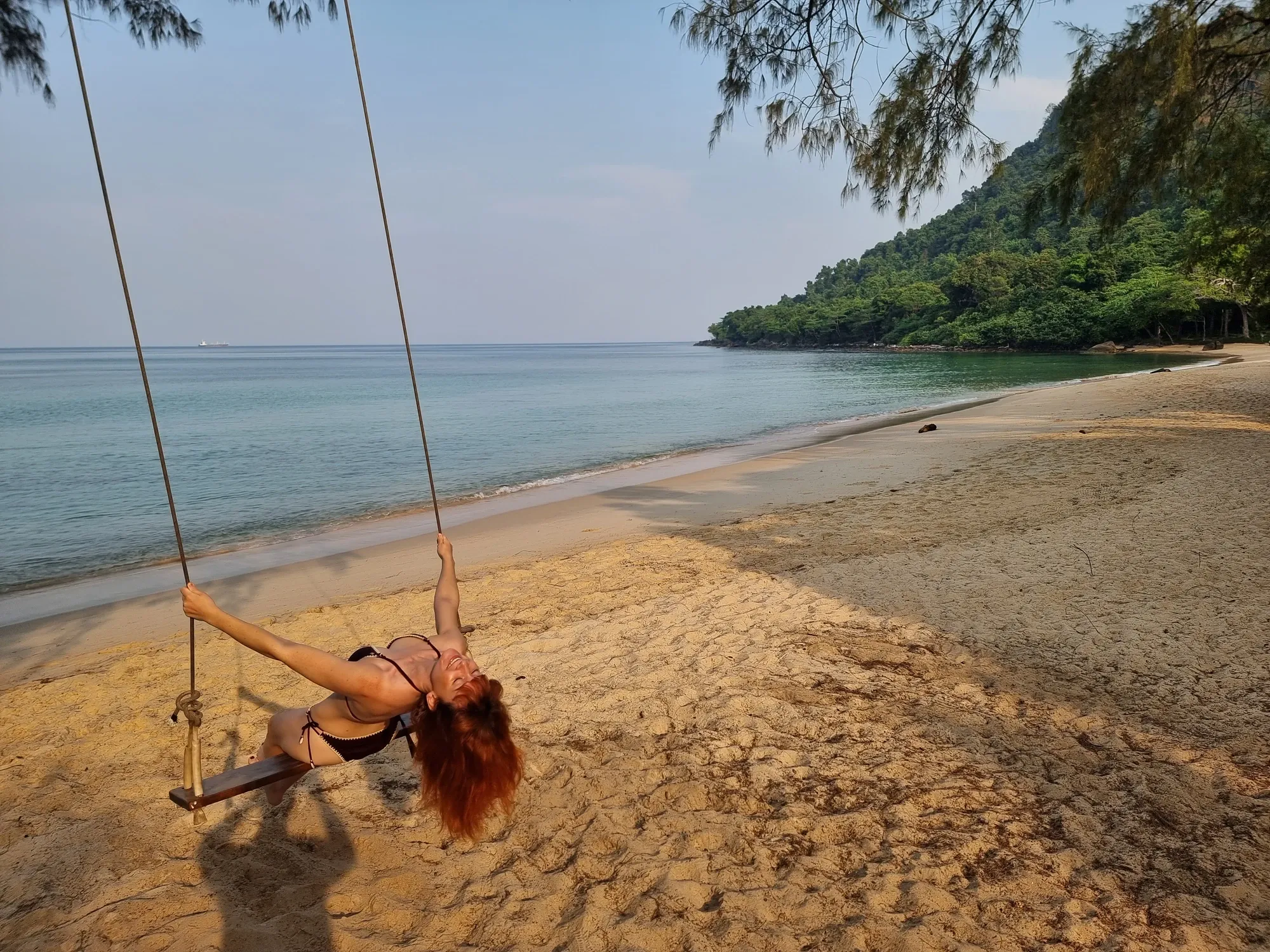 Ségolène se balance, la tête renversée, sur une balançoire suspendue au-dessus du sable de Sunset Beach, déserte au matin, face à la mer et à la côte boisée de Koh Rong Sanloem