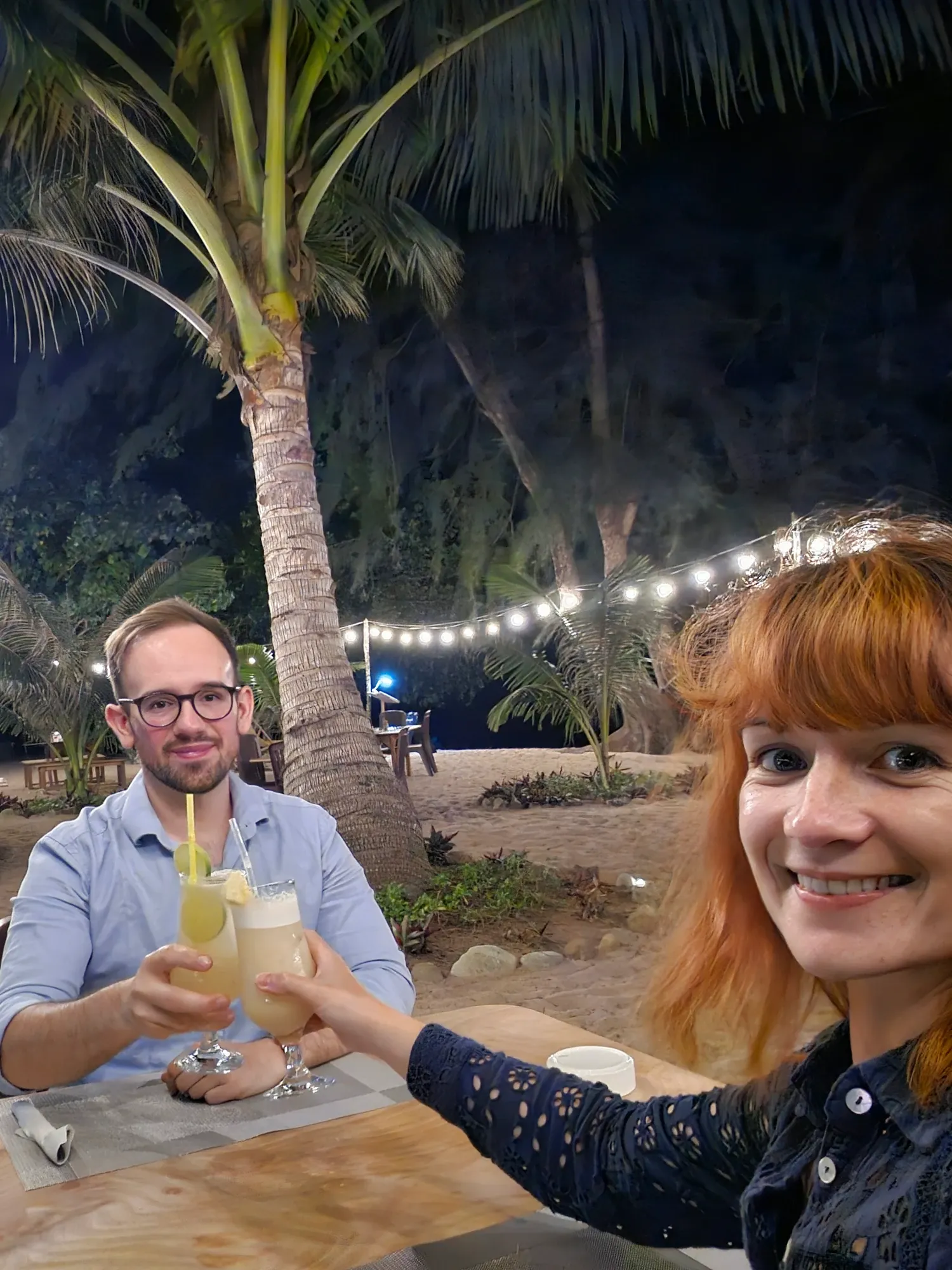 Antoine et Ségolène trinquent avec deux cocktails, de nuit, à une table posée sur le sable de Sunset Beach, sous un palmier et une guirlande lumineuse