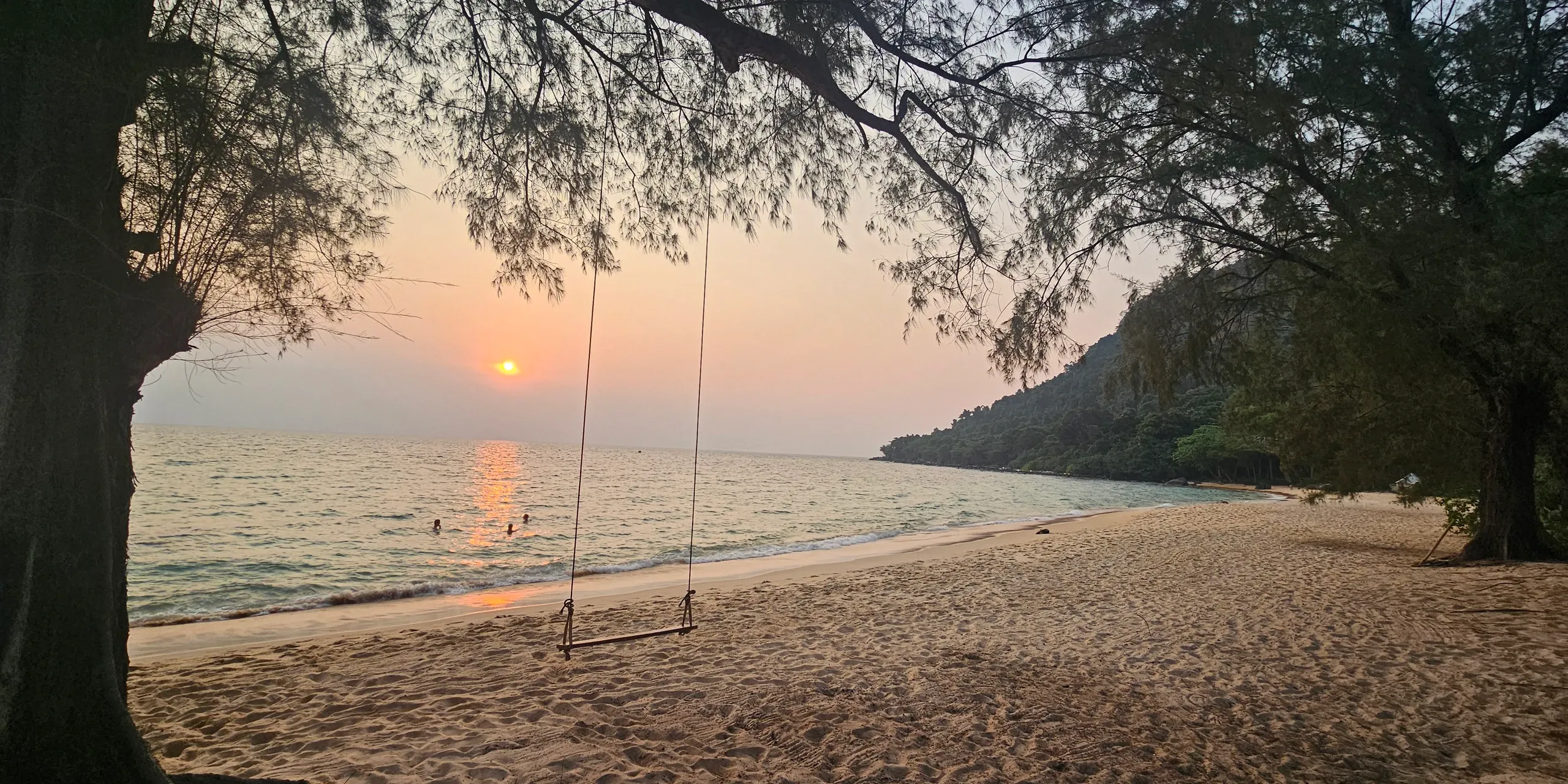 Coucher de soleil sur Sunset Beach, à Koh Rong Sanloem : une balançoire en bois pend d'un arbre au-dessus du sable, face à la mer où se baignent quelques personnes, avec la côte boisée de l'île à droite