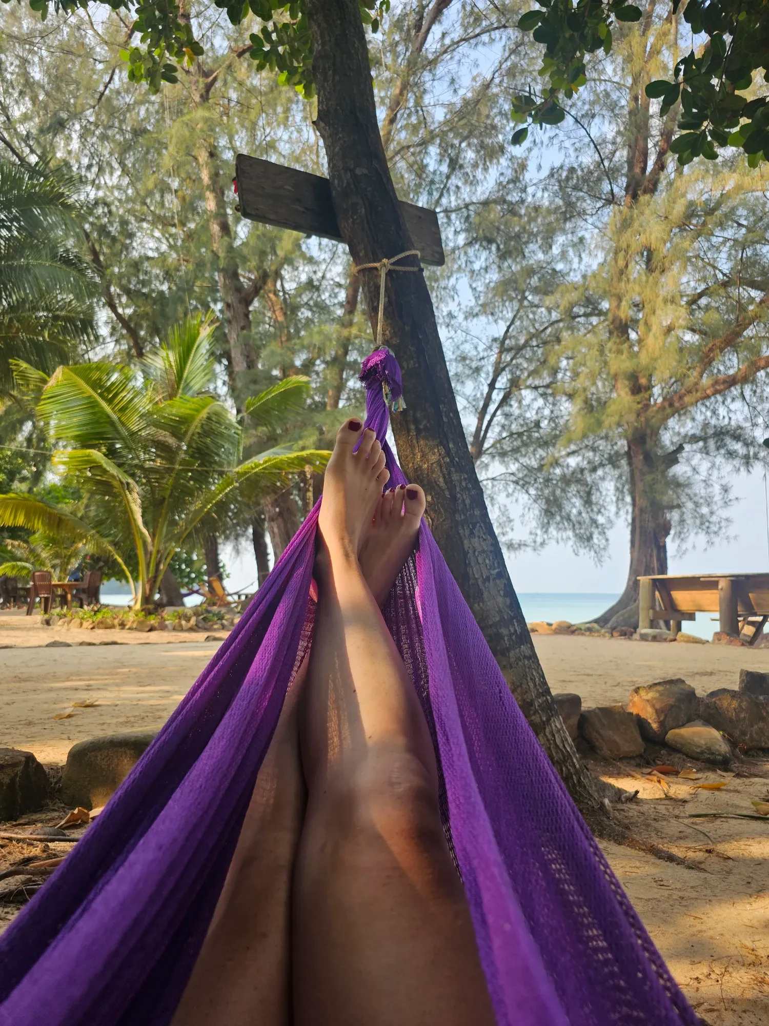 Jambes croisées dans un hamac violet accroché à un arbre, face au sable de Sunset Beach, à ses arbres et à la mer turquoise au loin