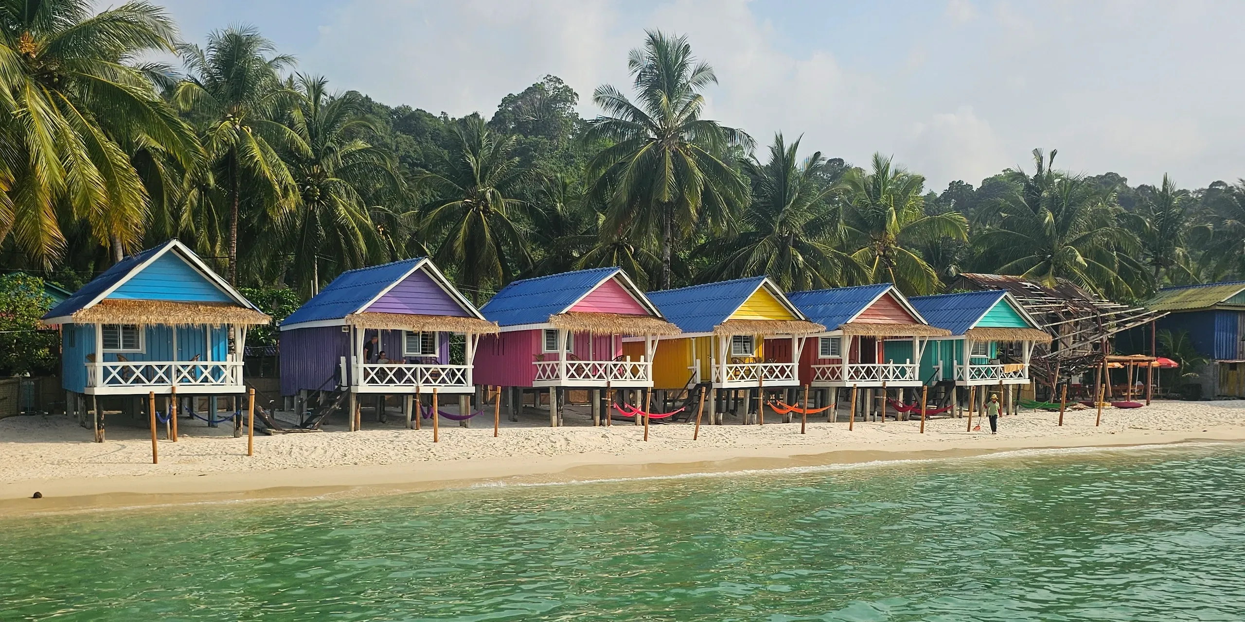 Rangée de bungalows sur pilotis aux façades colorées, bleu, violet, rose, jaune, orange et turquoise, sur une plage de sable blanc bordée de palmiers, à Sok San, vue depuis la mer turquoise