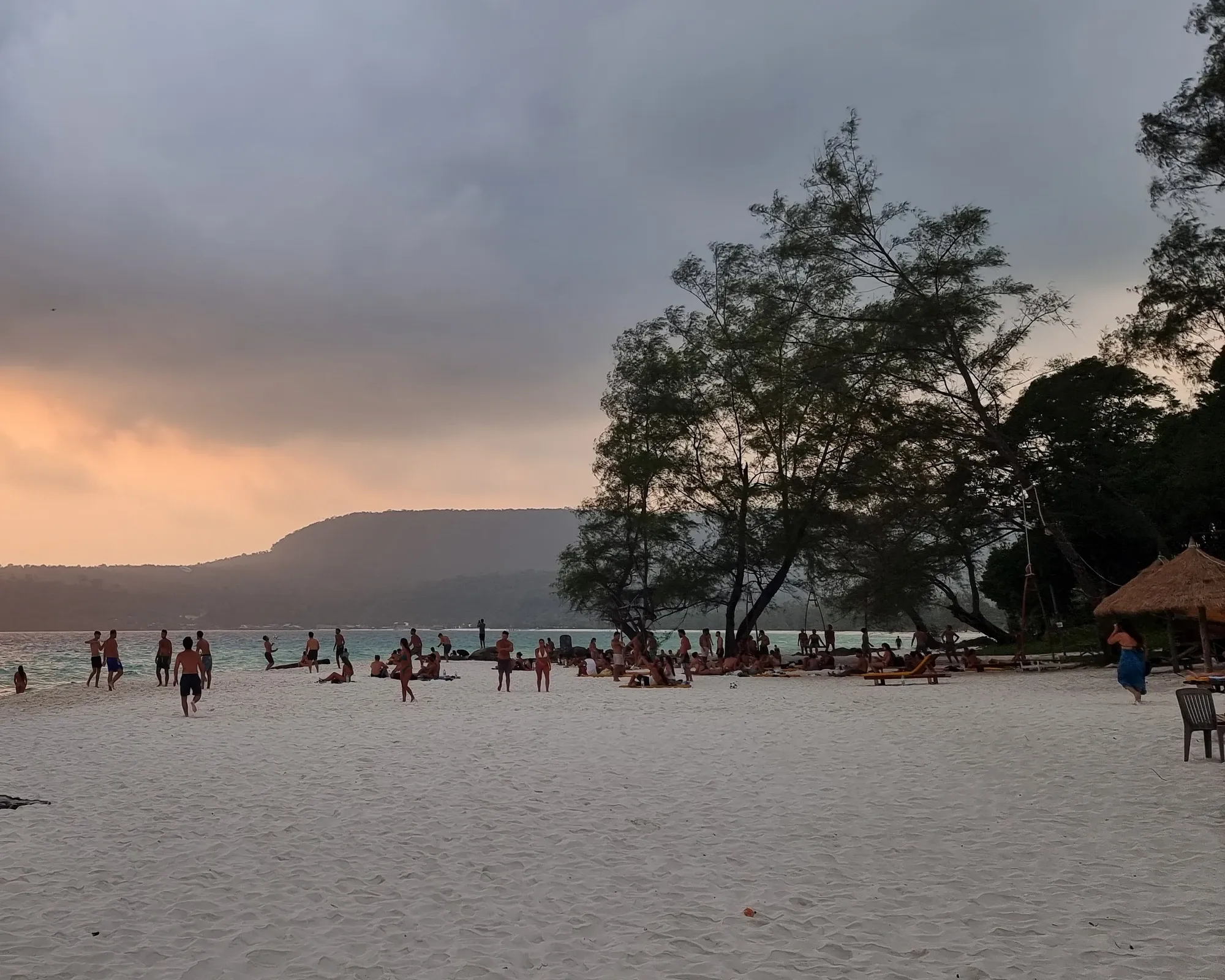 Plage de sable blanc animée au coucher du soleil, avec de nombreux baigneurs sous de grands arbres, une paillote à droite et une colline à l'horizon, sous un ciel chargé
