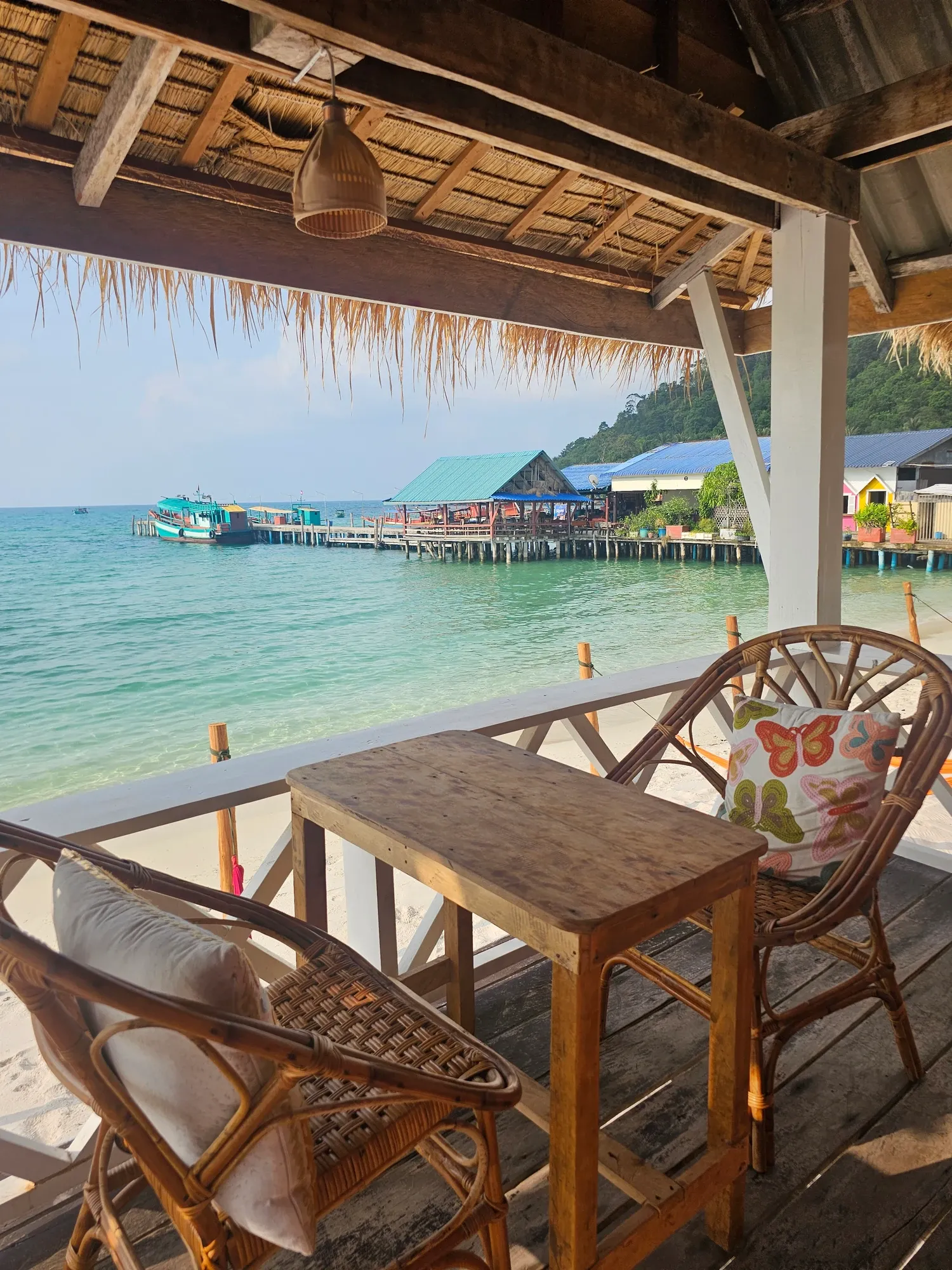 Terrasse de bungalow avec deux fauteuils en rotin et une petite table en bois, face à l'eau turquoise et à un embarcadère sur pilotis où sont amarrés des bateaux