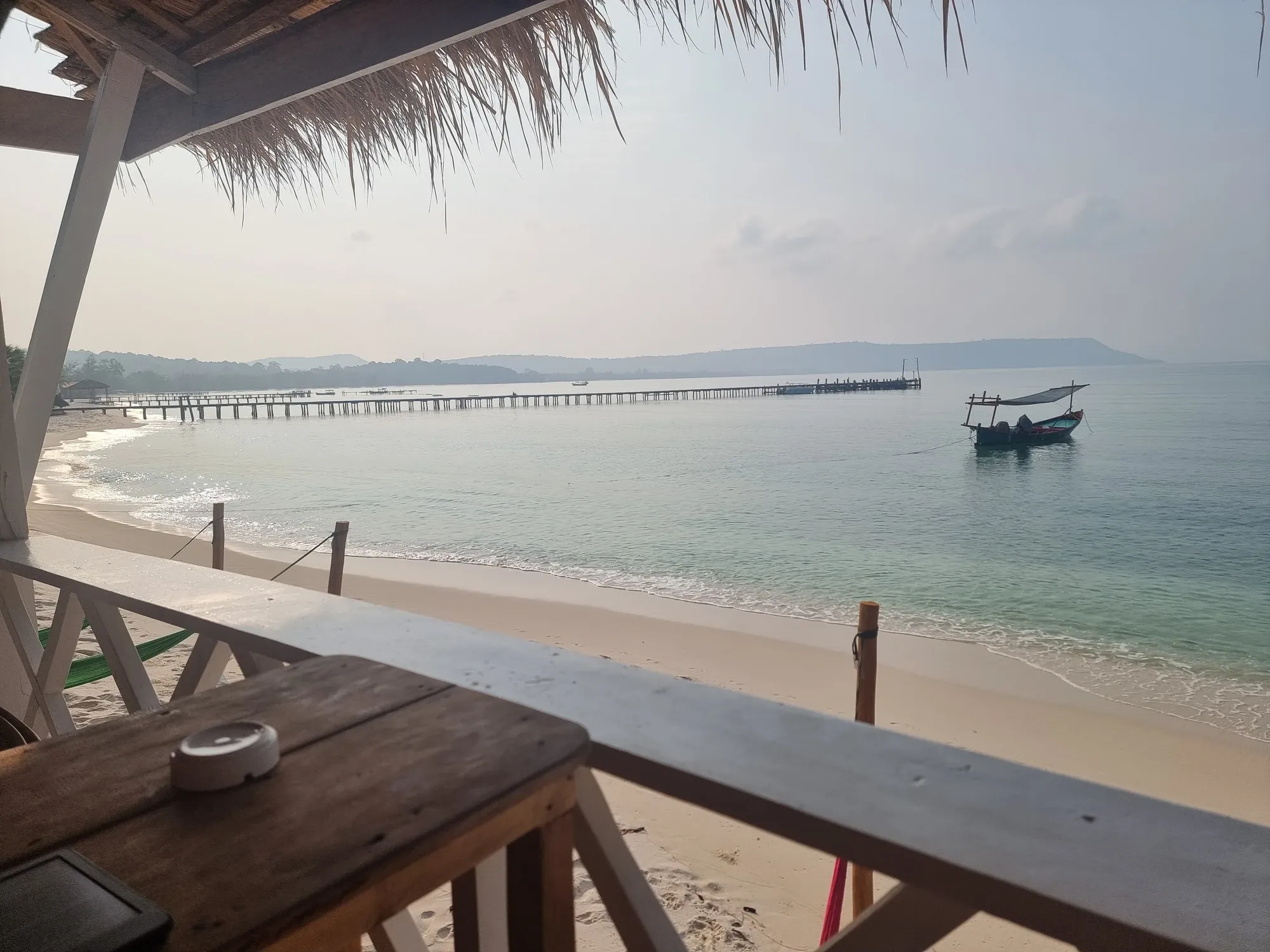 Vue depuis une terrasse sous un toit de chaume : la plage, un long ponton en bois qui s'avance dans la mer calme et un bateau de pêche au mouillage