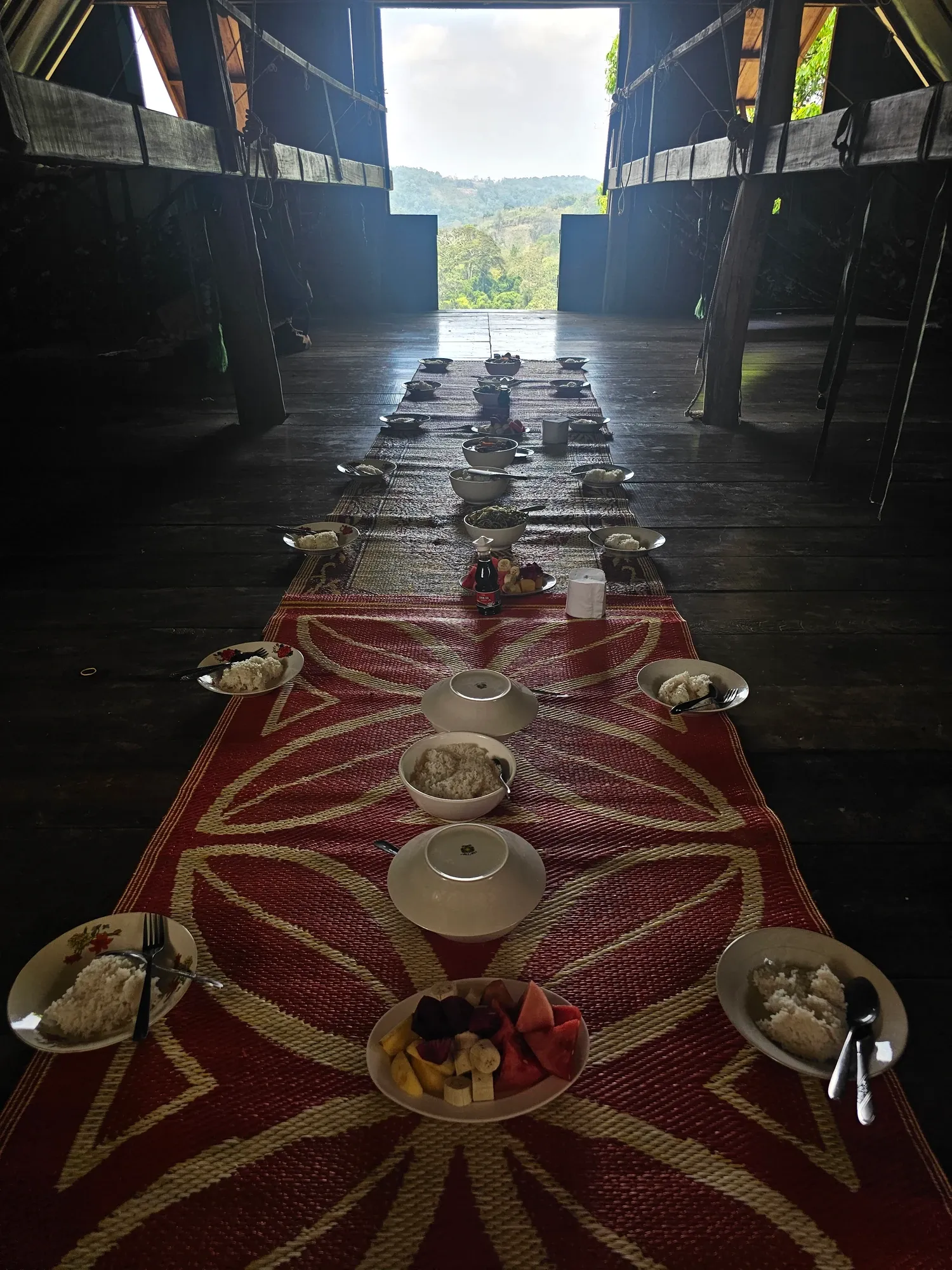 Un repas servi au sol sur une natte rouge dans une grande salle en bois sombre, avec du riz, des plats et des fruits, face à une ouverture sur les collines