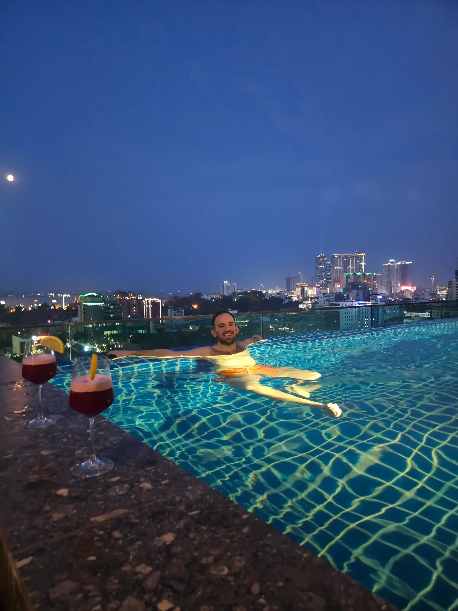 Antoine dans la piscine sur le toit du CheckInn Royal Palace, deux cocktails au bord, Phnom Penh illuminée au fond