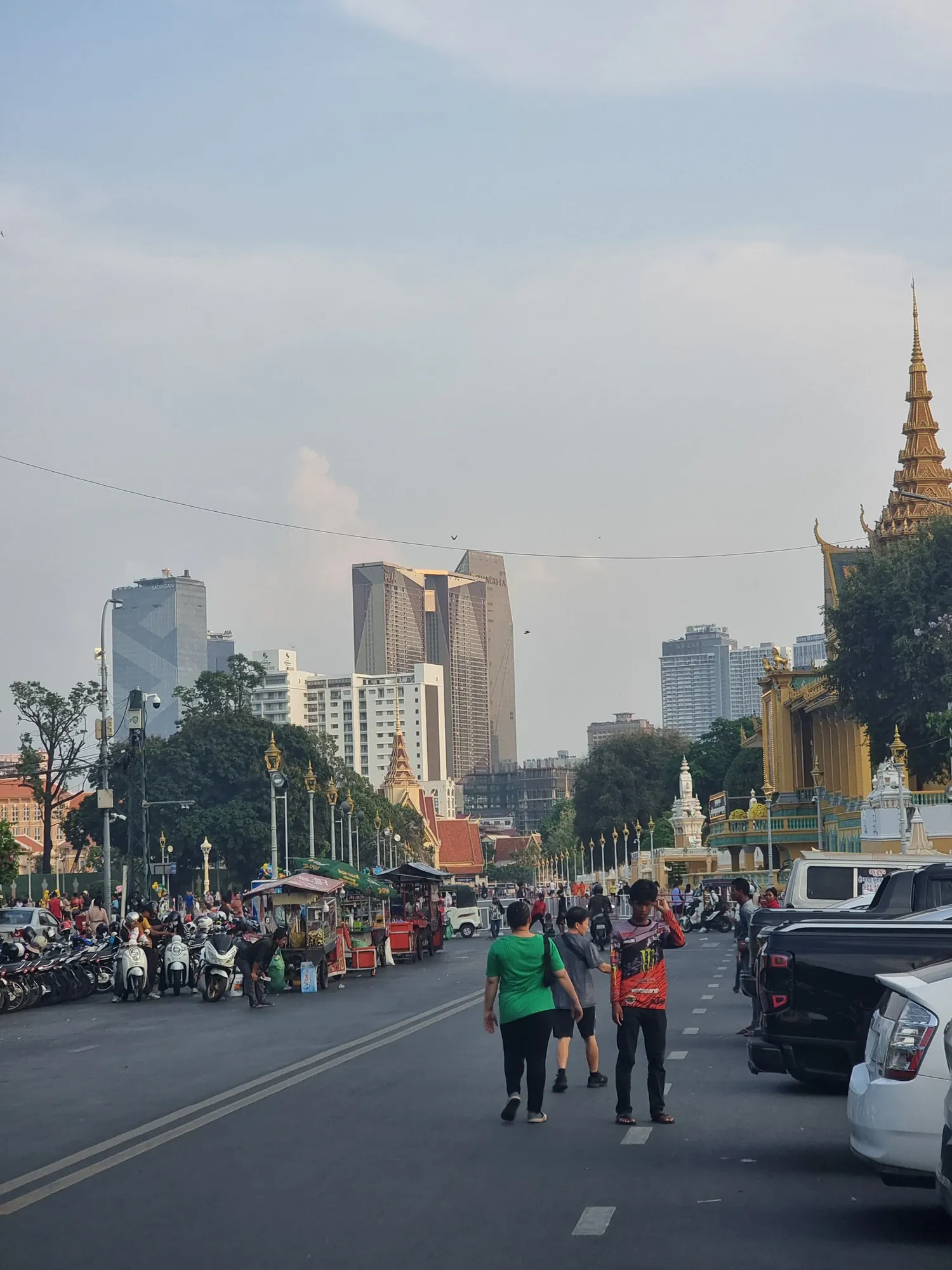 La rue devant le Palais Royal de Phnom Penh, avec ses étals, ses scooters garés et les tours du centre-ville au fond