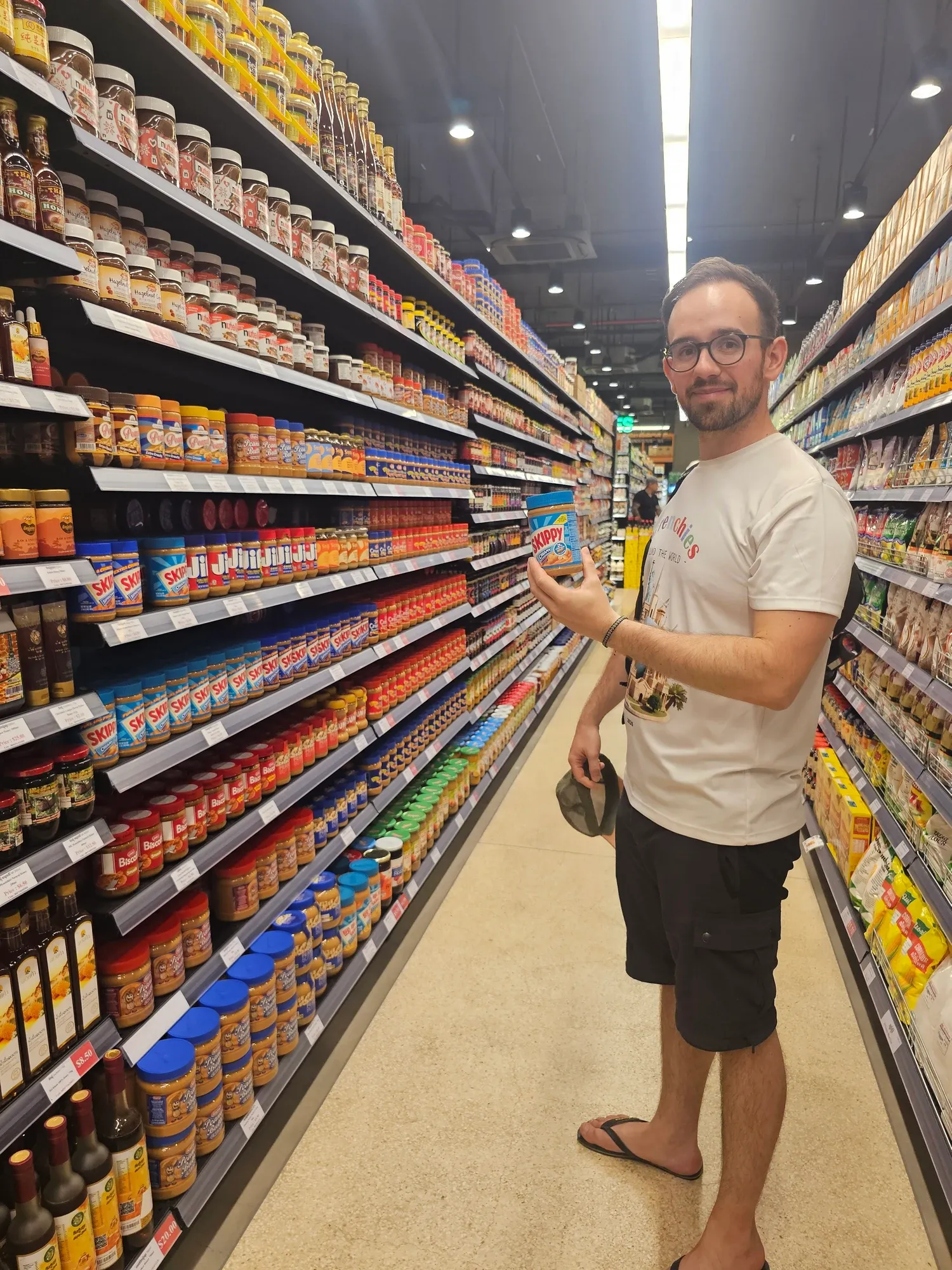 Antoine tient un pot de beurre de cacahuète Skippy devant un rayon entier de pots de beurre de cacahuète, au supermarché Angkor Market II