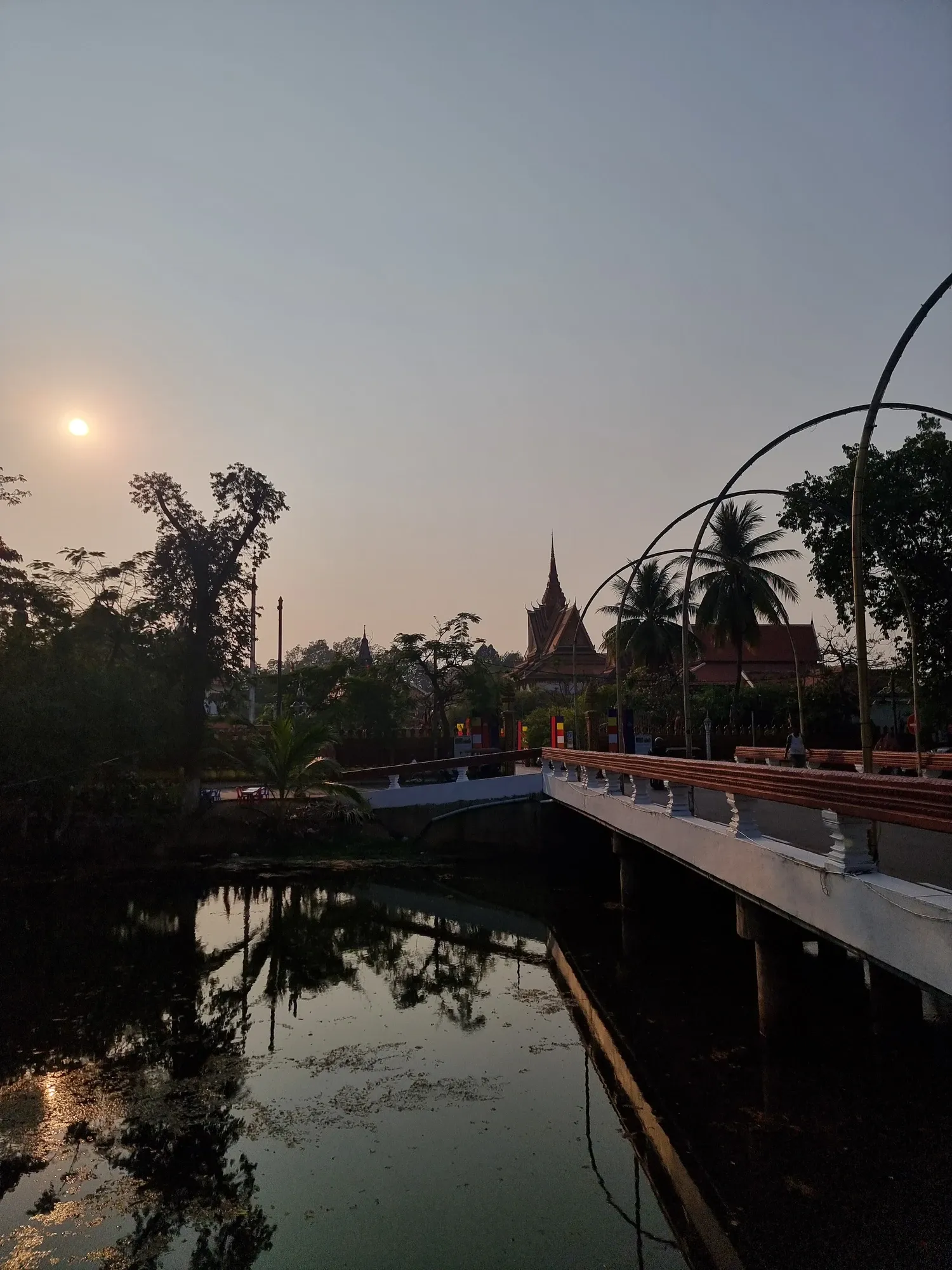 Coucher de soleil voilé sur un plan d'eau de Siem Reap, avec un pont aux rambardes ocre, des palmiers et le toit pointu d'une pagode à l'horizon